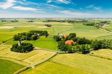 Luftaufnahme eines Traktors beim Mähen des Grases auf einem Acker mit Bauernhäusern und einer kleinen Kirche in Friesland, Niederlande. - AAEF09922