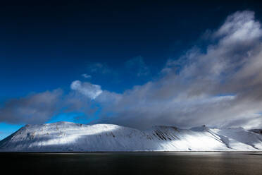 Atemberaubender Blick auf einen wolkenverhangenen Himmel über einem verschneiten Bergkamm und einem ruhigen See an einem Wintertag in Island - ADSF27336