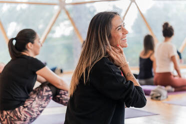 Seitenansicht einer lächelnden langhaarigen Frau in schwarzen langen Ärmeln, die ihren Hals berührt und in einem hellen Raum beim Yoga wegschaut - ADSF27367