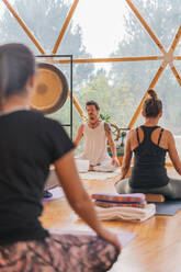 Back view of relaxed balanced group of people sitting with crossed leg and listening trainers decorated class for yoga with transparent walls - ADSF27384