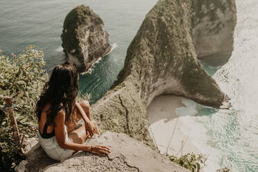 Rückenansicht einer jungen Frau mit langen Haaren, die auf einem hohen Felsen sitzt und aus der Höhe auf eine malerische Meereslandschaft blickt - ADSF27415