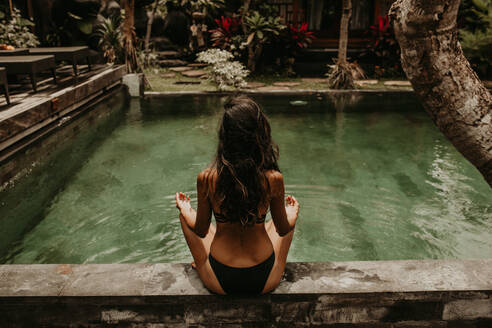 Back view of slim female in bikini practicing yoga on poolside of tropical spa hotel, Bali - ADSF27417