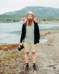 Attraktiver bärtiger Hipster in Freizeitkleidung, der eine professionelle Fotokamera hält und an der Küste in der Nähe von Wasser steht - ADSF27434