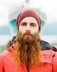 Gutaussehender bärtiger Reisender mit Hut und warmer Jacke, der in die Kamera schaut, während er vor dem unscharfen Hintergrund eines Schiffs im Hafen steht - ADSF27438