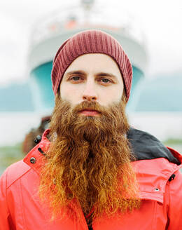 Handsome bearded traveler in hat and warm jacket looking at camera while standing on blurred background of ship in harbor - ADSF27438