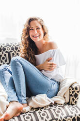 Beautiful cheerful young woman looking at camera sitting on armchair in room - ADSF27459