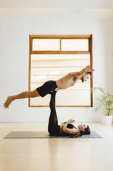 Mann über Frau, die eine Gleichgewichtsposition in einer Yoga-Sitzung im Trainingsraum einnimmt - ADSF27495
