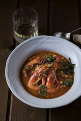 From above of delicious spicy stew with chickpeas and big shrimps decorated with cilantro leaves served in white soup plate on wooden table with glass of drink and spoon - ADSF27537