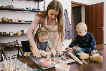 Ein Kind sitzt am Tisch, während ein Handwerksmeister und ein Mädchen in einer hellen Werkstatt Ton mit einem Nudelholz rollen - ADSF27563