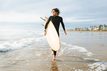 Surfer in Neoprenanzug geht mit Surfbrett in Richtung Wasser, um eine Welle am Strand bei Sonnenaufgang zu erwischen - ADSF27573