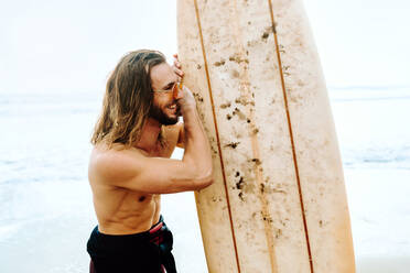 Junger, fröhlicher, gut aussehender Surfer mit langen Haaren, Neoprenanzug und stylischer Sonnenbrille, der mit seinem Surfbrett in Richtung Wasser schaut, um eine Welle am Strand zu erwischen - ADSF27575