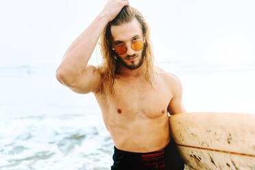 Junger Surfer mit langen Haaren, Neoprenanzug und stylischer Sonnenbrille, der mit seinem Surfbrett in Richtung Wasser schaut, um eine Welle am Strand zu erwischen - ADSF27577