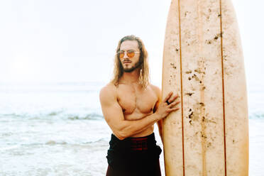 Junger Surfer mit langen Haaren, Neoprenanzug und stylischer Sonnenbrille, der mit seinem Surfbrett in Richtung Wasser schaut, um eine Welle am Strand zu erwischen - ADSF27578