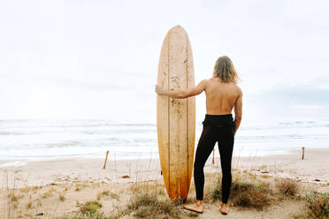 Rückenansicht eines nicht erkennbaren Surfers mit langen Haaren in einem Neoprenanzug, der mit einem Surfbrett in einer Sanddüne steht - ADSF27579