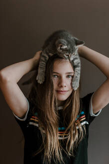 Dreamy teenage girl with fluffy cute cat on head on brown background in studio - ADSF27610