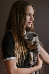 Dreamy thoughtful teenage girl holding fluffy cute cat on brown background in studio - ADSF27611