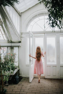 Back view of unrecognizable female with long ginger hair opening door of greenhouse with plants - ADSF27612