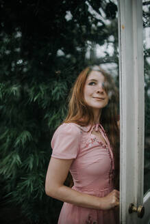 Verträumte positive rothaarige Frau im Kleid steht in der Nähe der Glastür eines Gewächshauses mit üppigen Pflanzen - ADSF27613