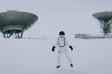 Unrecognizable cosmonaut in spacesuit standing in snowy valley in winter on background of huge radar antennas in Svalbard - ADSF27620