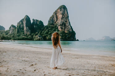 Rückansicht Ganzkörperansicht einer gesichtslosen Frau im weißen Kleid am Sandstrand in der Nähe von azurblauem Wasser gegen felsige Klippen in Thailand - ADSF27626