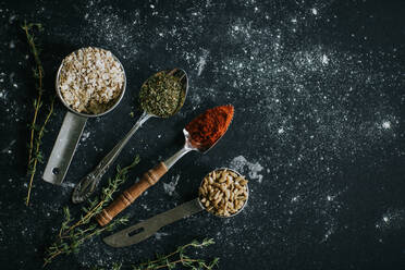 Top view of spoons with paprika and dry herbs placed on black table with oat and sunflower seeds - ADSF27629