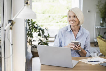 Lächelnde Geschäftsfrau, die wegschaut und eine Tasse im Büro zu Hause hält - UUF24010