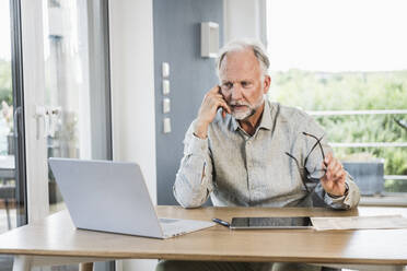 Businessman with eyeglasses thinking while looking at laptop in home office - UUF24027