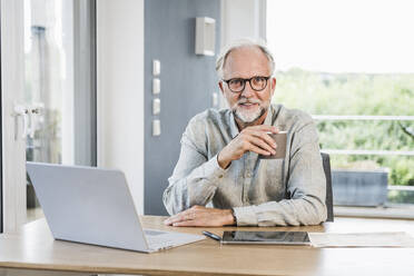 Geschäftsmann mit Brille hält Tasse am Schreibtisch im Home Office - UUF24028