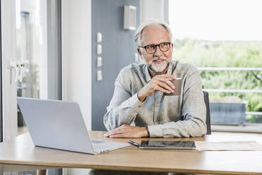 Businessman looking away holding while coffee mug at desk in home office - UUF24029