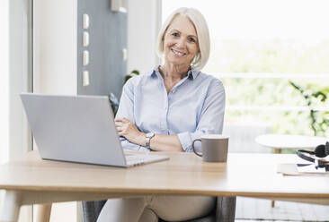 Mature female professional with arms crossed sitting at desk in home office - UUF24042