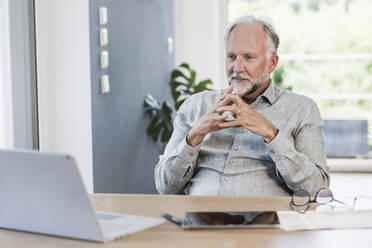 Businessman with hands clasped looking at laptop on desk in home office - UUF24044