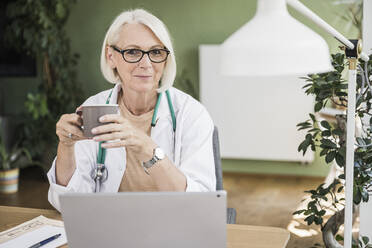 Ältere Ärztin hält Kaffeetasse am Schreibtisch - UUF24047