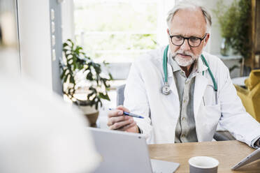 Mature male doctor looking at laptop while holding pen at desk - UUF24054