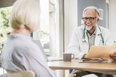 Lächelnder männlicher Arzt im Gespräch mit einer Patientin am Schreibtisch - UUF24058