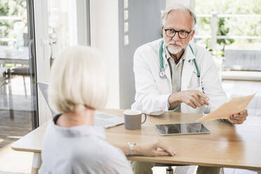 Männlicher Arzt hält einen medizinischen Bericht und betrachtet eine Patientin am Schreibtisch - UUF24061