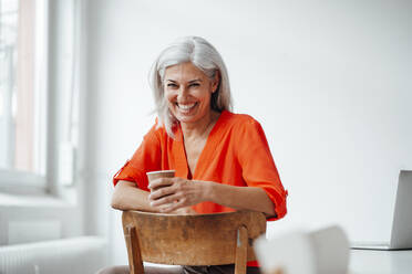 Reife Geschäftsfrau mit weißem Haar, die auf einem Stuhl im Büro Kaffee trinkt - JOSEF05080
