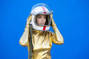 Young woman looking away while wearing space helmet in front of blue wall - DAMF00852