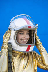 Junge Frau lächelt, während sie einen Weltraumhelm vor einer blauen Wand trägt - DAMF00853