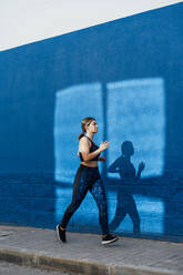 Young woman jogging on footpath by blue wall - DAMF00859