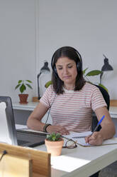 Female customer service representative writing on diary while talking through headphones at office - VEGF04712