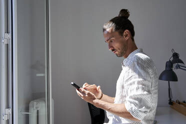 Männlicher Berufstätiger, der ein Mobiltelefon vor einem Fenster im Büro benutzt - VEGF04730