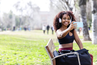 Smiling female athlete with headphones taking selfie through smart phone at park - RFTF00099