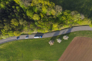 Drohnenansicht von Autos, die auf einer Landstraße fahren, die sich zwischen einem grünen Hain und einem gepflügten Feld erstreckt - RUEF03355