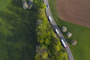 Drohnenansicht von Lastwagen, die auf einer Landstraße fahren, die sich zwischen einem grünen Hain und einem gepflügten Feld erstreckt - RUEF03356