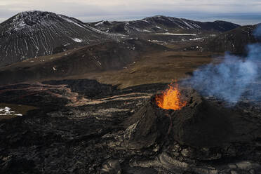 Luftaufnahme eines aus dem Krater rauchenden Vulkans, Blick auf aus dem Krater ausbrechendes Magma mit Berglandschaft im Hintergrund, Grindav√≠k, Südliche Halbinsel, Island. - AAEF10336
