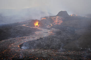 Luftaufnahme von Lava, die durch die schmelzenden Felsen auf dem Berggipfel fließt, mit Magma, das aus Kratern spritzt, und Rauch ringsum, M√Ωrdalshreppur, Island. - AAEF10354