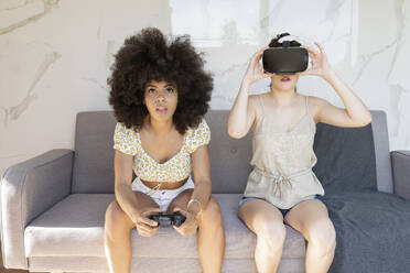 Afro-Frau spielt ein Videospiel, während ihre Freundin zu Hause ein Virtual-Reality-Headset trägt - JCCMF03234