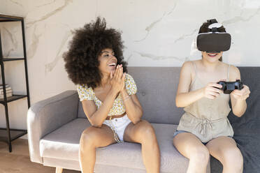 Afro-Frau gestikuliert, während sie eine Freundin beim Spielen mit einem Virtual-Reality-Headset zu Hause beobachtet - JCCMF03237