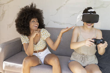 Young woman playing game while female friend gesturing sitting on sofa at home - JCCMF03238