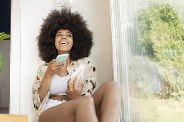 Lächelnde Frau, die ein Buch hält, während sie eine Sprachnachricht über ein Smartphone zu Hause sendet - JCCMF03255